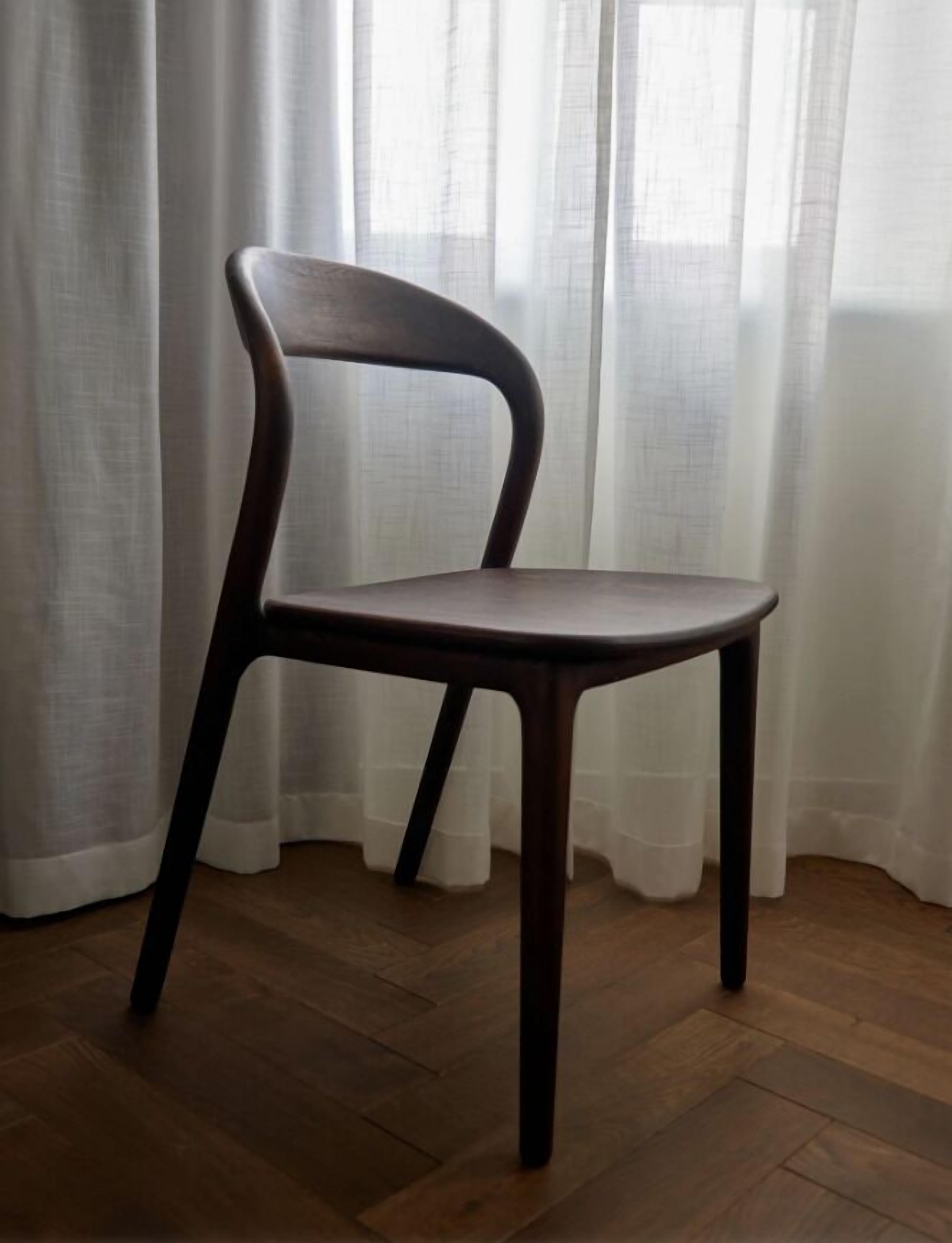 Una silla de madera con respaldo curvo y cuatro patas, ubicada frente a una ventana con cortinas traslúcidas.