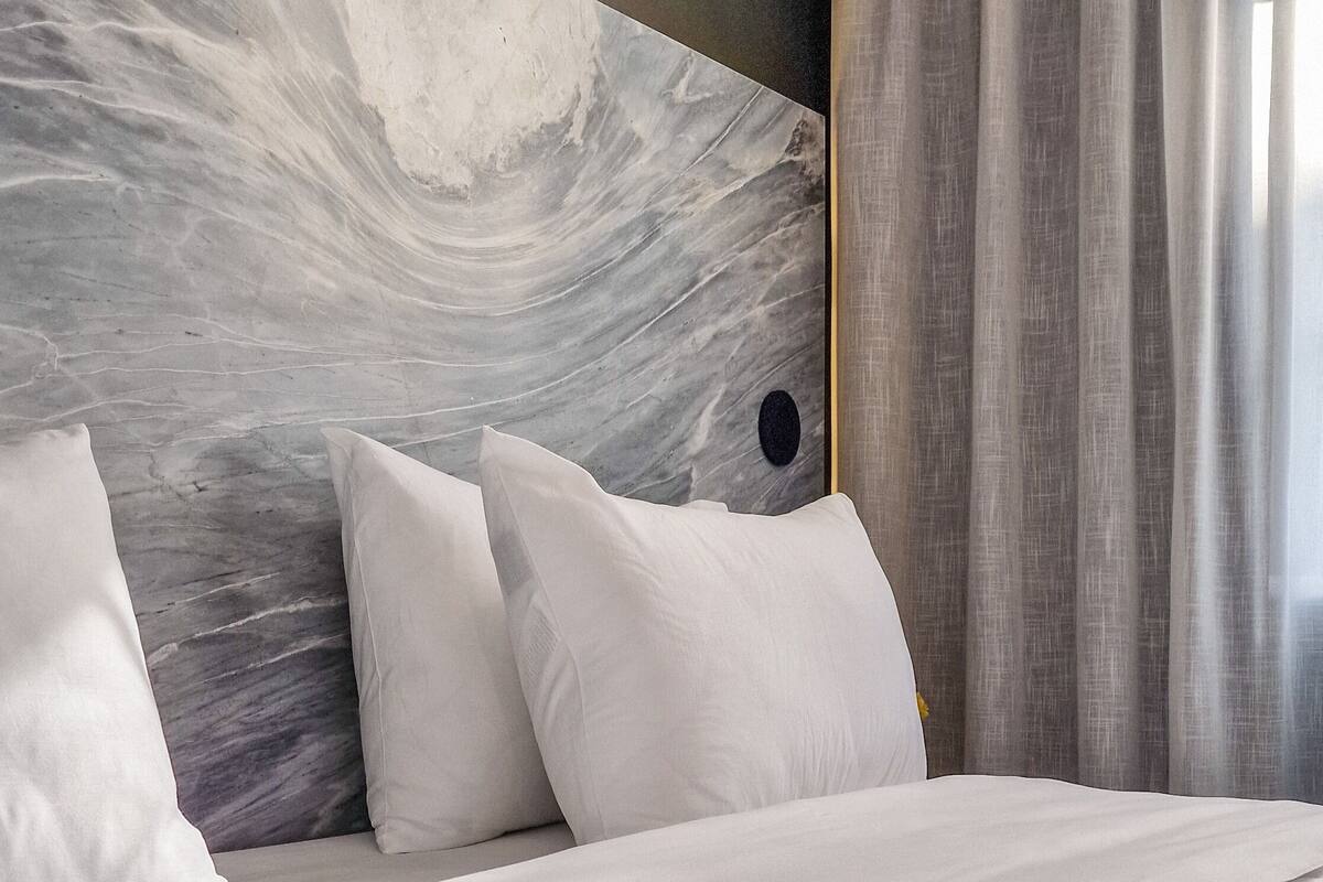 Una cama impecablemente tendida con sábanas y almohadas blancas, un cabecero de mármol y cortinas translúcidas.