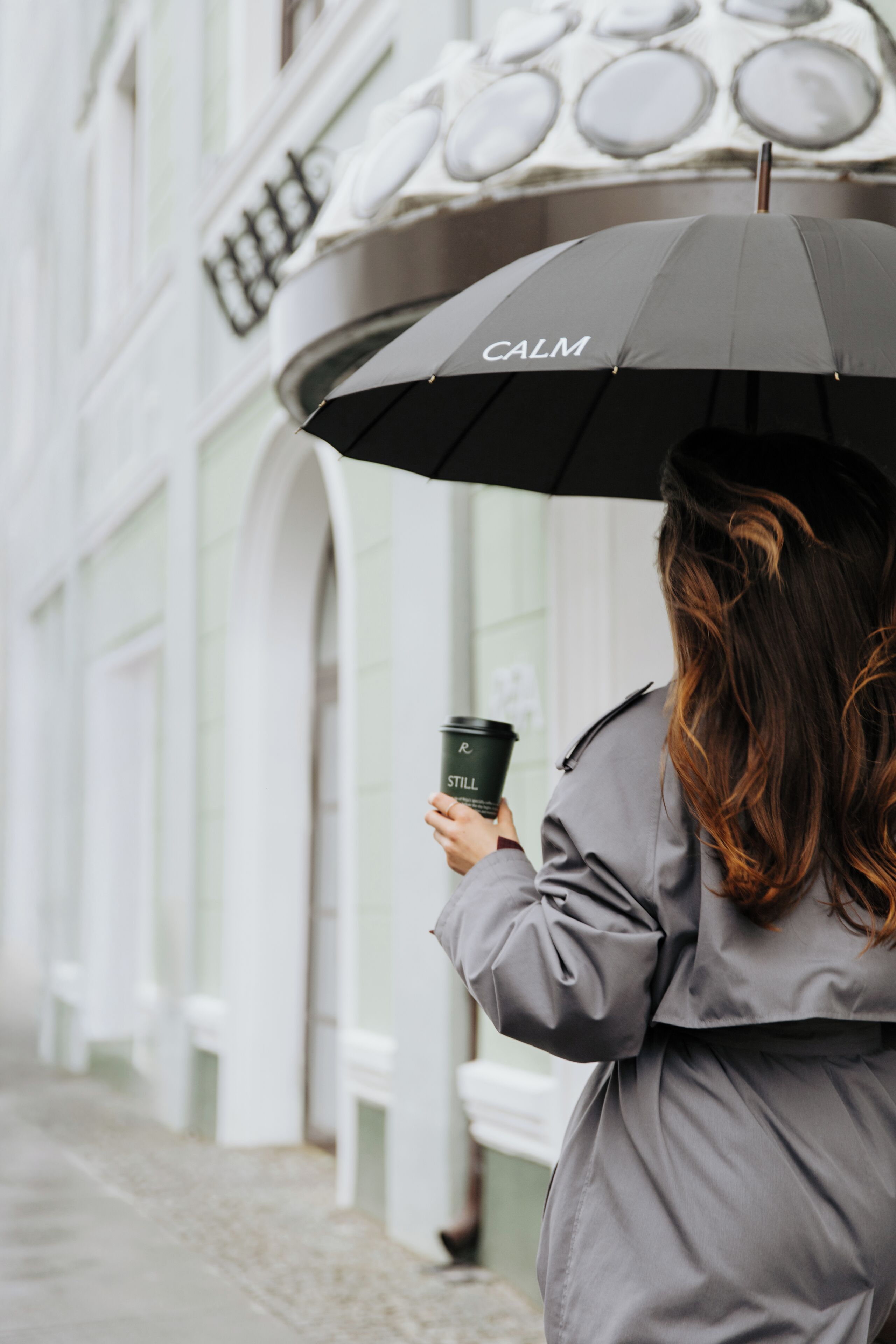 Une personne tenant un parapluie noir sur lequel est écrit le mot « CALME », marchant sur une rue pavée.