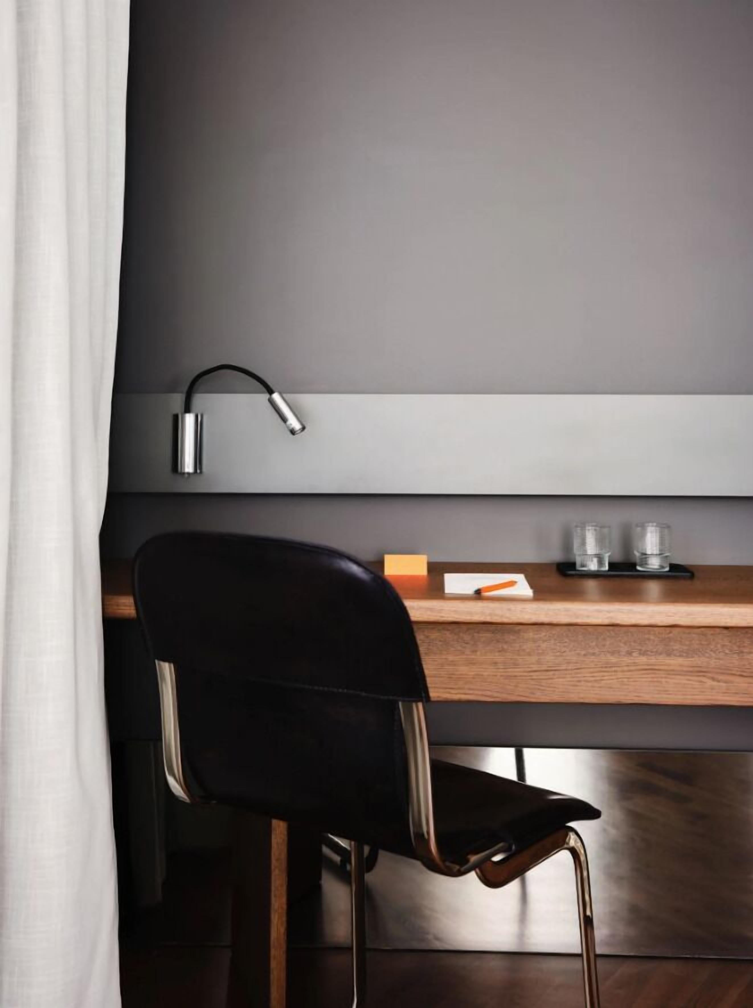 Una habitación moderna con un escritorio de madera, una silla negra, una lámpara de escritorio y un estante pequeño con dos vasos.