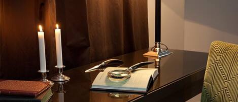 A wooden table with two lit candles, a green lamp, a book, and a magnifying glass.