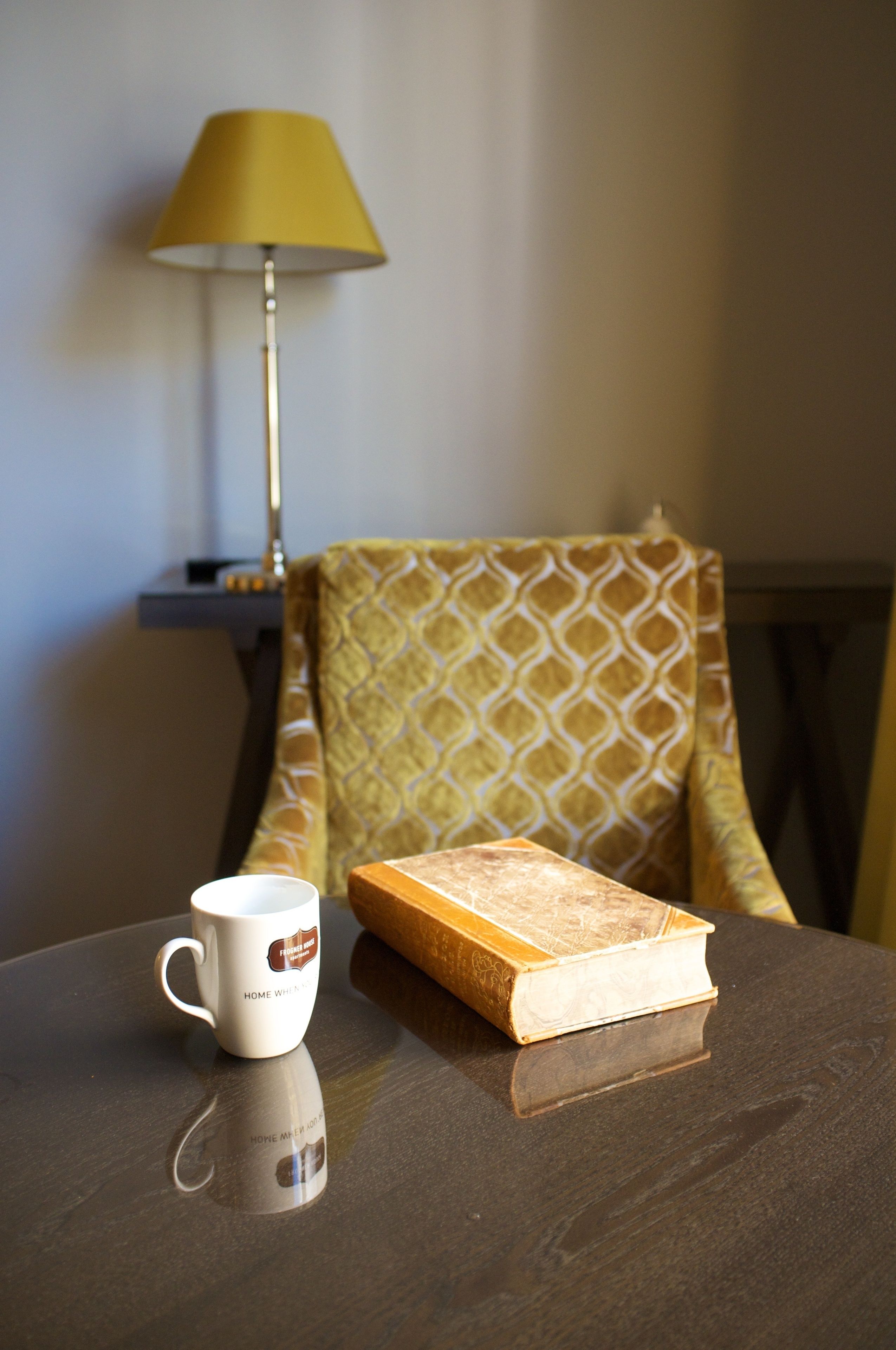 Una mesa con una taza blanca, un libro, una lámpara y una silla estampada.