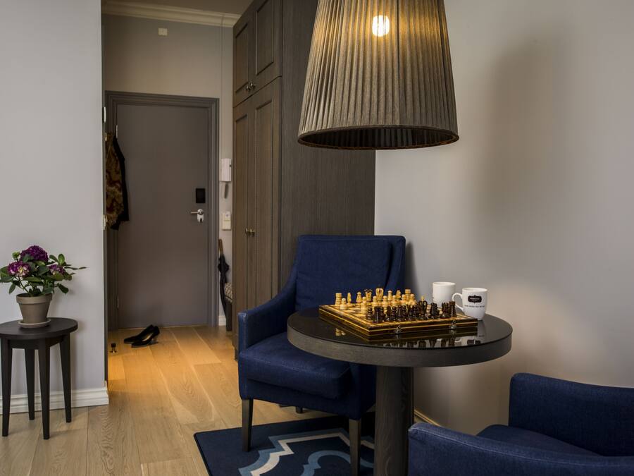 Una habitación con un sillón azul, una mesa redonda con un tablero de ajedrez, una planta en maceta y una lámpara colgante.