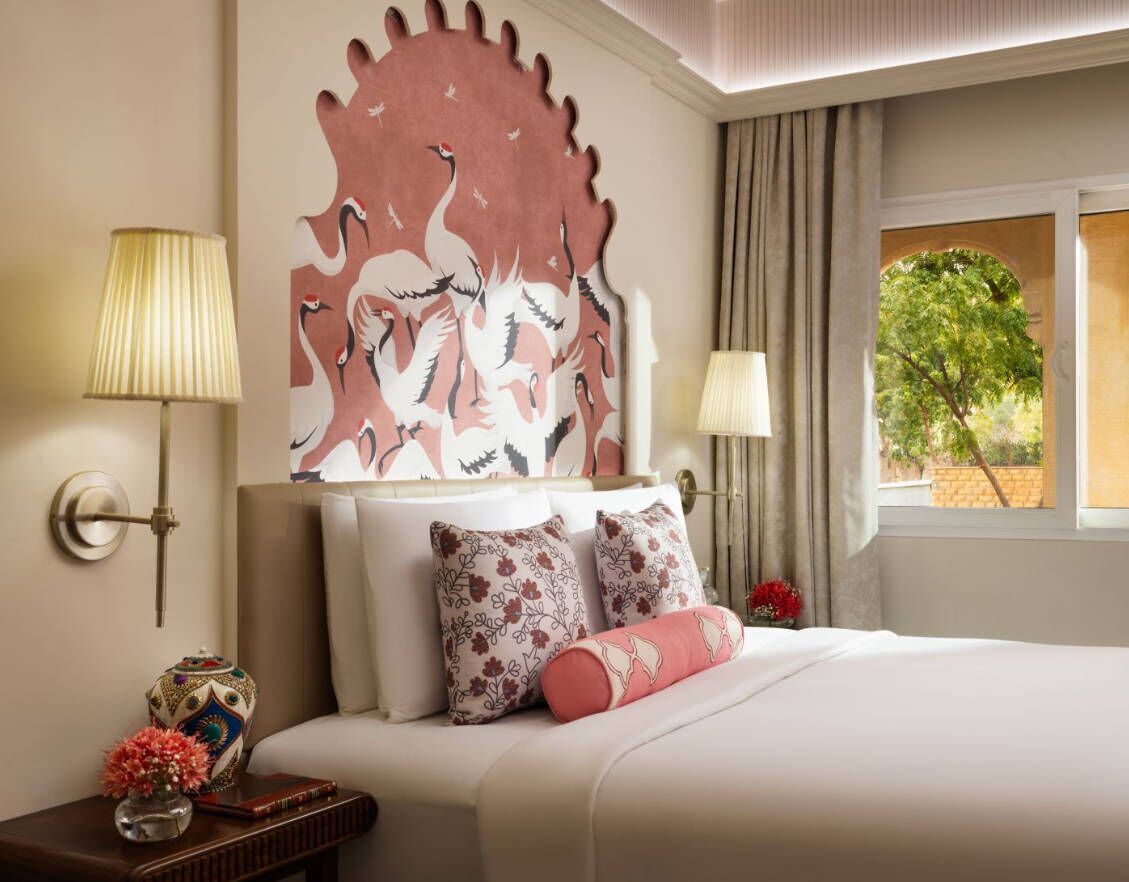 Une chambre d’hôtel avec une grande fresque murale, un lit agrémenté d’oreillers à motifs floraux, une table de chevet avec un vase et un livre, et une fenêtre donnant sur un paysage verdoyant.