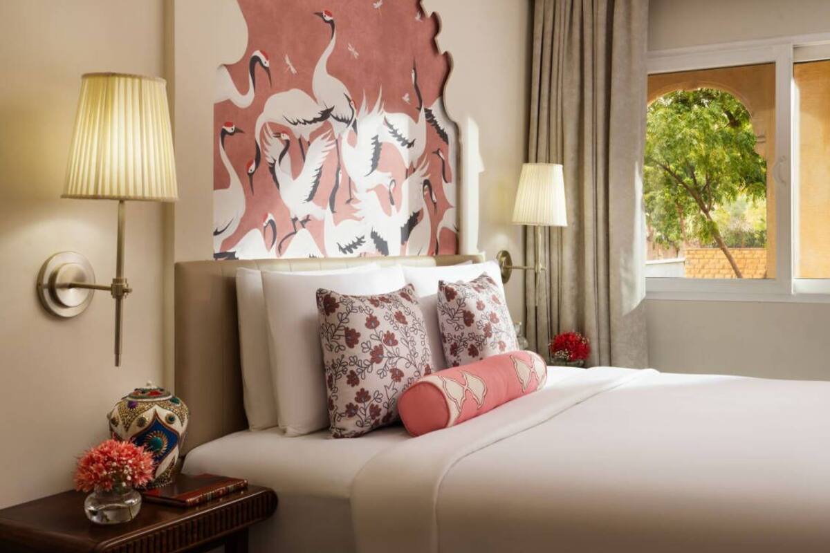 Une chambre d’hôtel avec une grande fresque murale, un lit agrémenté d’oreillers à motifs floraux, une table de chevet avec un vase et un livre, et une fenêtre donnant sur un paysage verdoyant.