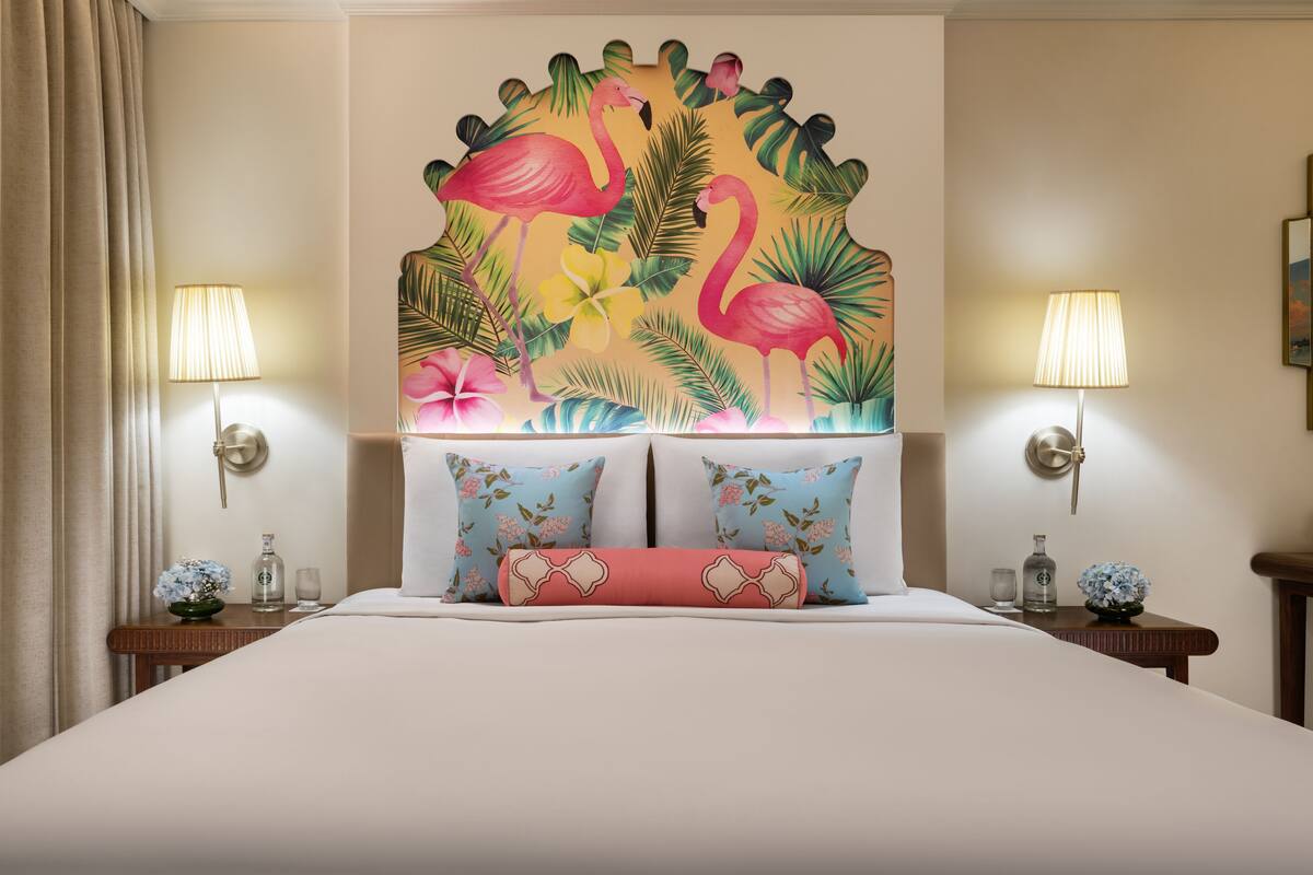 Une chambre d’hôtel avec une grande fresque murale sur le thème des tropiques, deux lampes de chevet, un lit avec des oreillers à motifs floraux et une table de chevet sur laquelle se trouvent un verre et une bouteille.