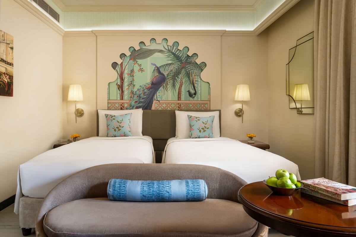 Une chambre d’hôtel avec deux lits, un canapé, une table avec un saladier de fruits et un livre, ainsi qu’une fresque murale décorative.