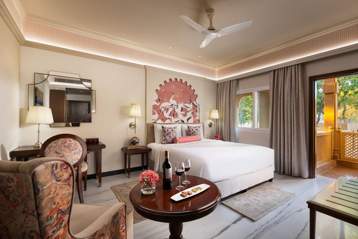 Une chambre d’hôtel avec un grand lit, un bureau, une chaise, une table avec une bouteille et des verres, et une vue sur l’extérieur.