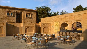 An outdoor seating area with wicker chairs and tables, set against a stone building with carved details and arched doorways. - Taj Gorbandh Palace, Jaisalmer (Jaisalmer)