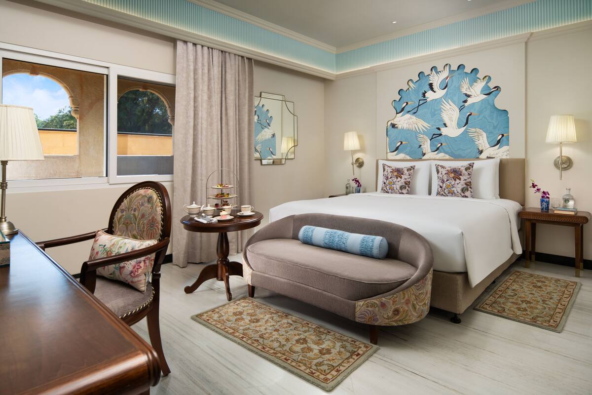 Une chambre d’hôtel avec un grand lit, un canapé, une table à manger et une chaise. Il y a deux lampes sur les tables de chevet et un élément décoratif mural au-dessus du lit.