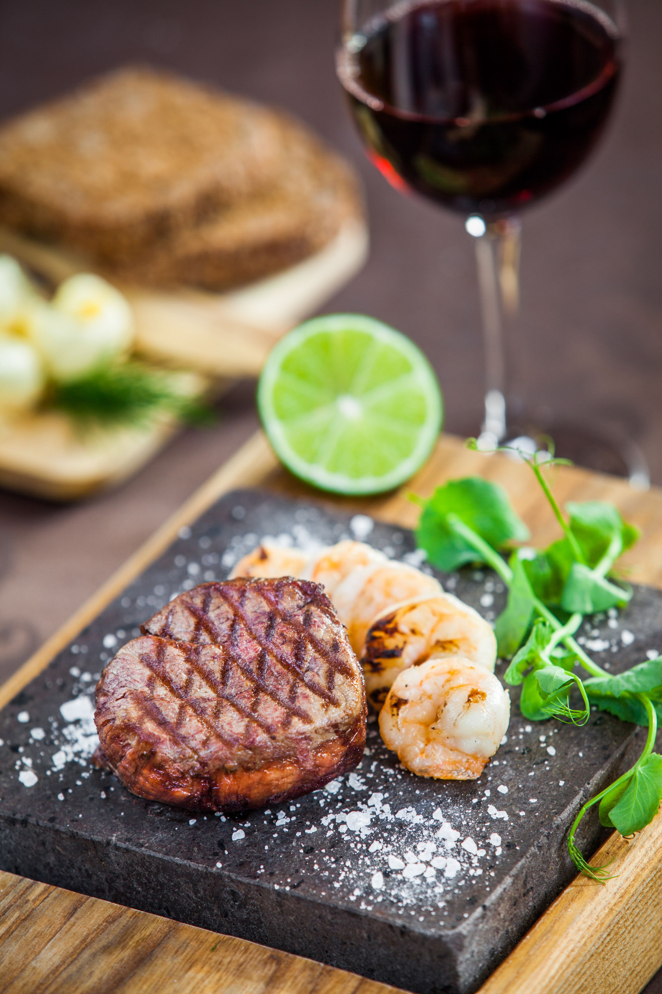 A steak with a grill mark, shrimp, a lime half, and a glass of red wine on a slate board.
