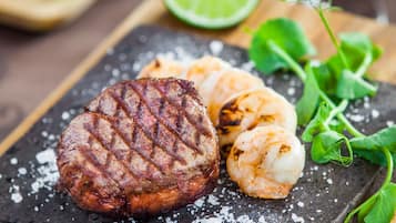 A steak with a grill mark, shrimp, a lime half, and a glass of red wine on a slate board.