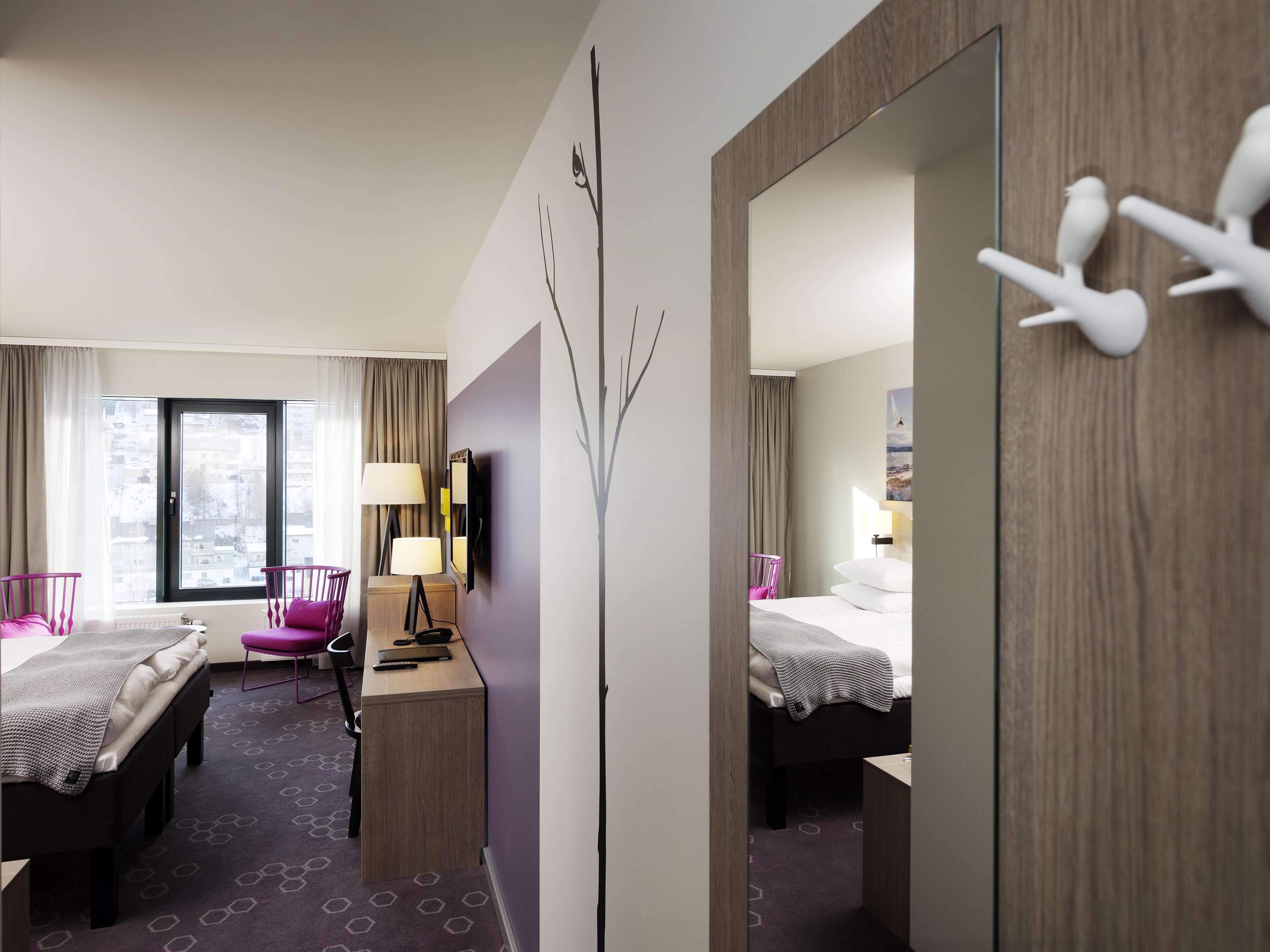 A hotel room with two beds, a desk, a chair, a mirror, and a view of the city.