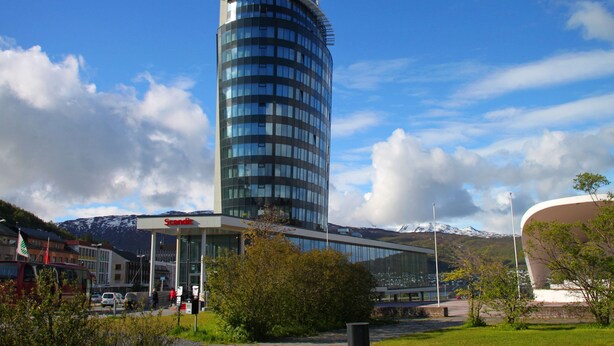 A modern hotel with a curved glass facade, a prominent 'Scandic' sign, and a view of a mountain range in the background.