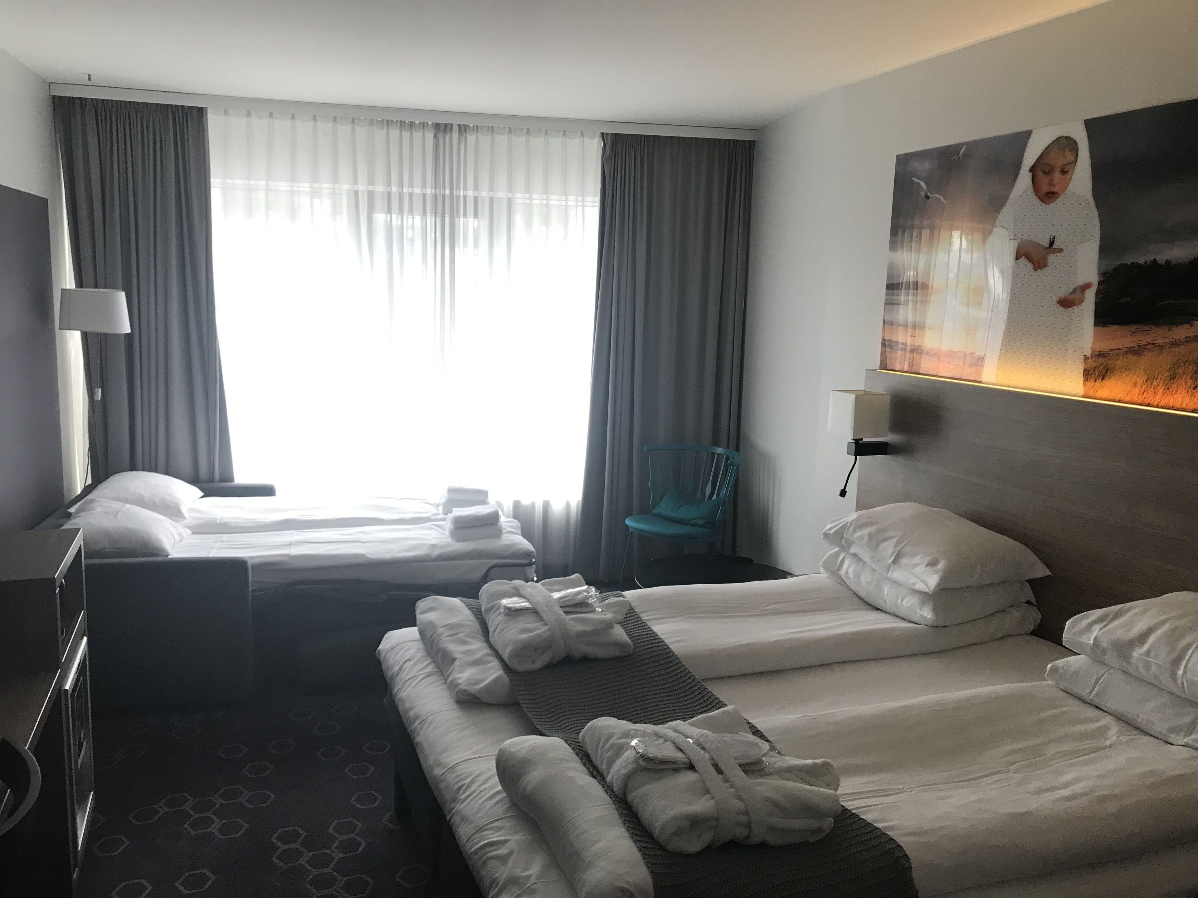 A hotel room with two beds, a chair, a lamp, and a painting on the wall.
