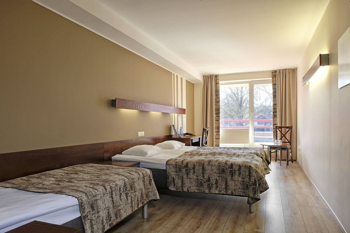 Une chambre d’hôtel avec deux lits, une tête de lit en bois, une petite table, une chaise et une fenêtre avec des rideaux.