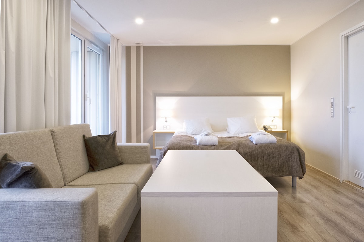 Une chambre d’hôtel moderne avec un grand lit, un canapé, une table de chevet et une grande fenêtre avec des rideaux.