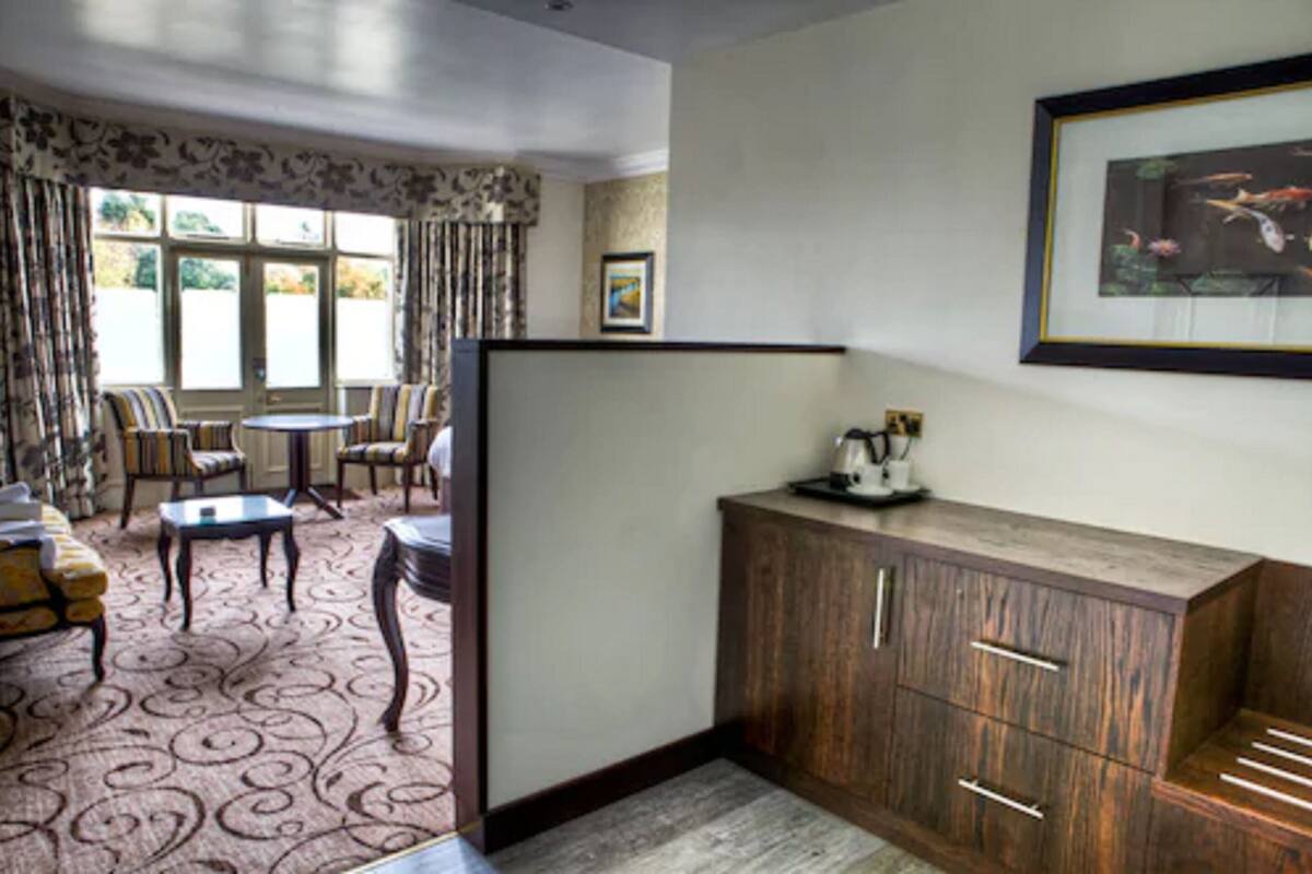 A room with a patterned carpet, a sofa, a dining table with chairs, a wooden cabinet, and a framed picture on the wall.