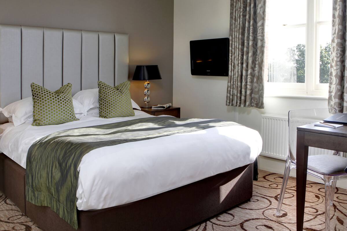 A hotel room with a large bed, a bedside table with a lamp, a TV mounted on the wall, and a window with curtains.