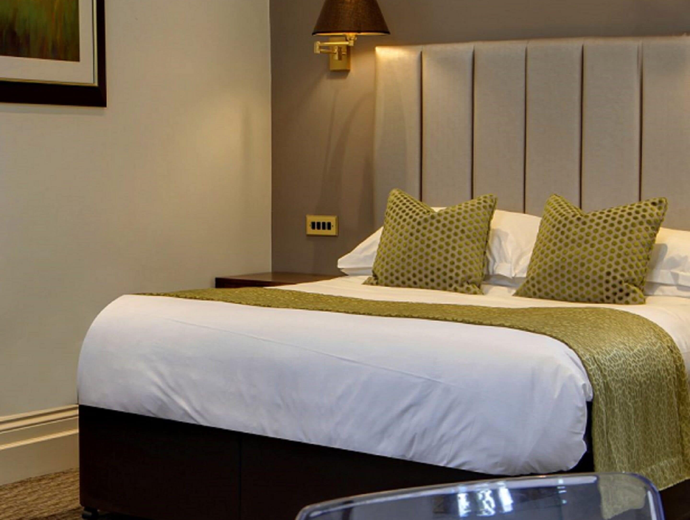 A neatly made bed with a white duvet and green accents, a wall-mounted lamp, and a headboard with vertical panels.