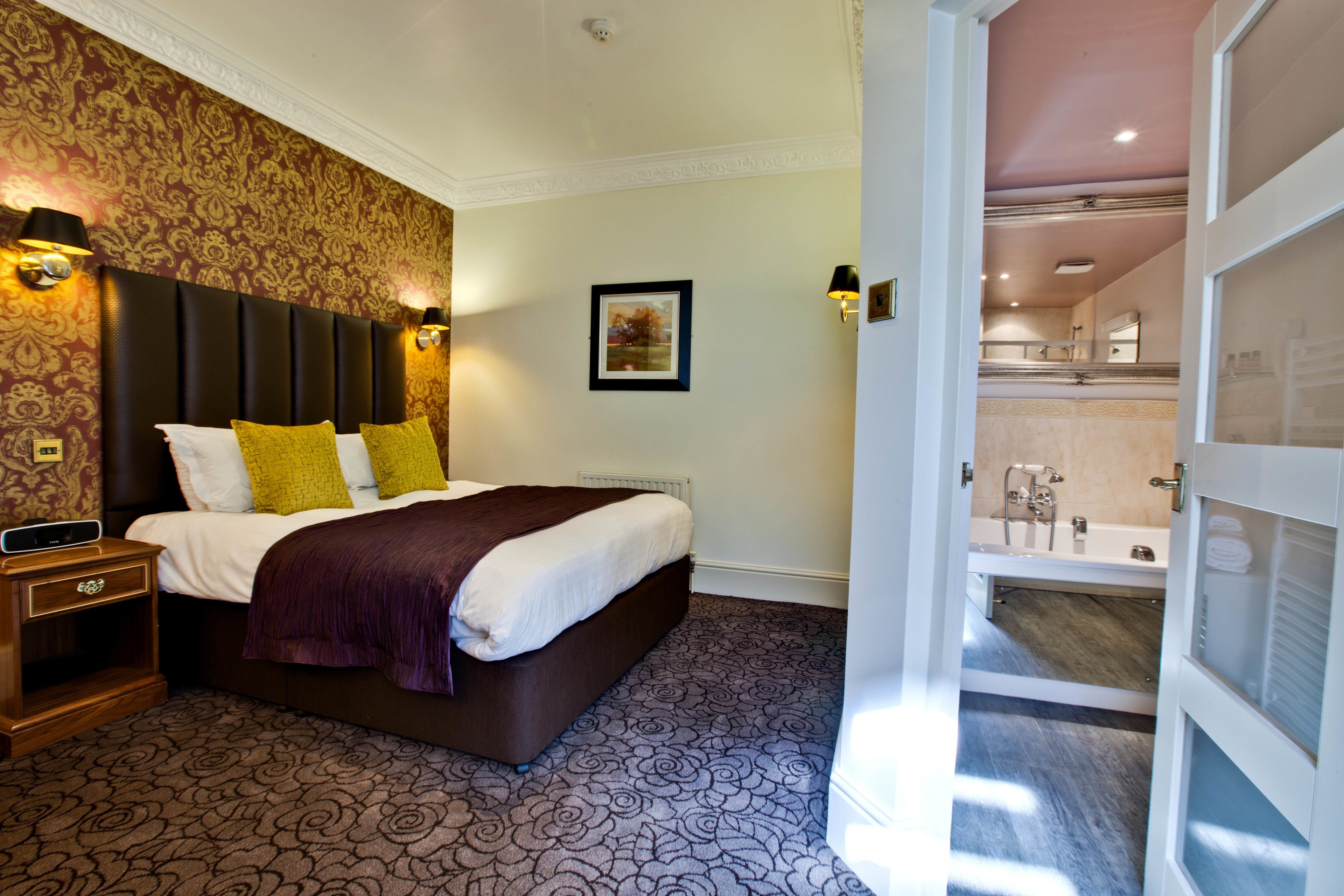 A hotel room with a large bed, a nightstand, a wall-mounted lamp, and a framed picture.