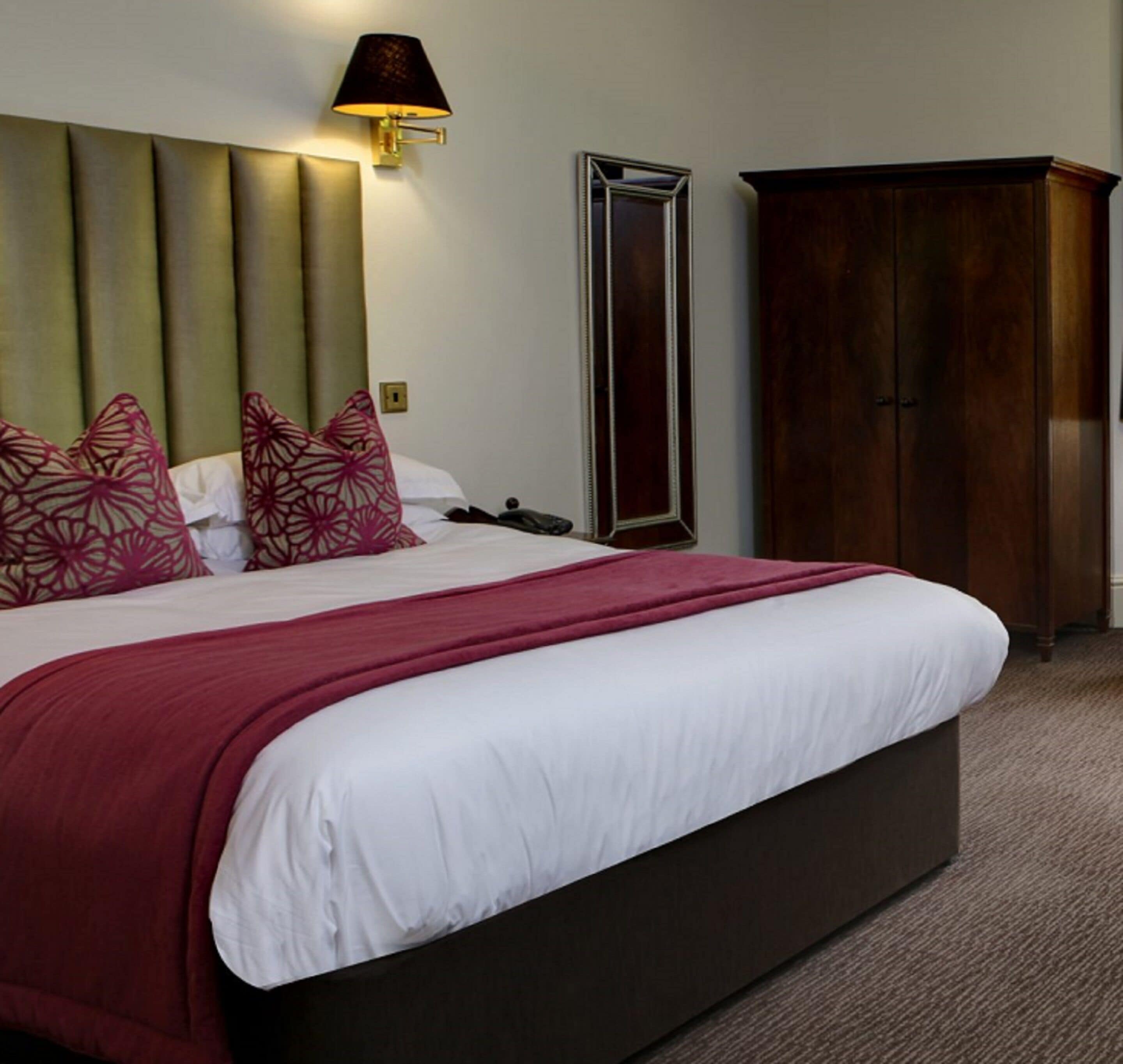 A hotel room with a large bed, a headboard, a bedside lamp, a mirror, and a wardrobe.