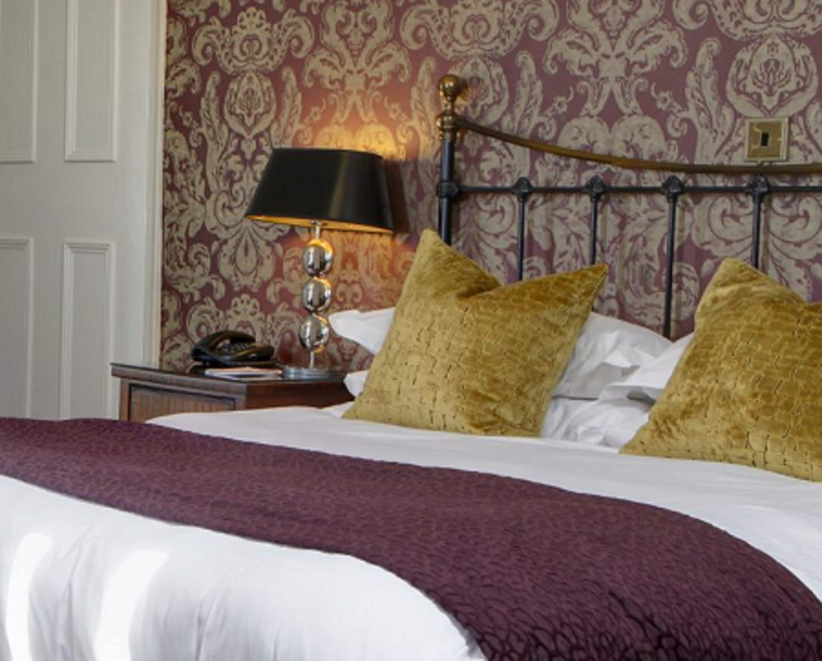 A neatly made bed with a purple blanket and two gold pillows, a bedside lamp, and a patterned wallpaper in a hotel room.
