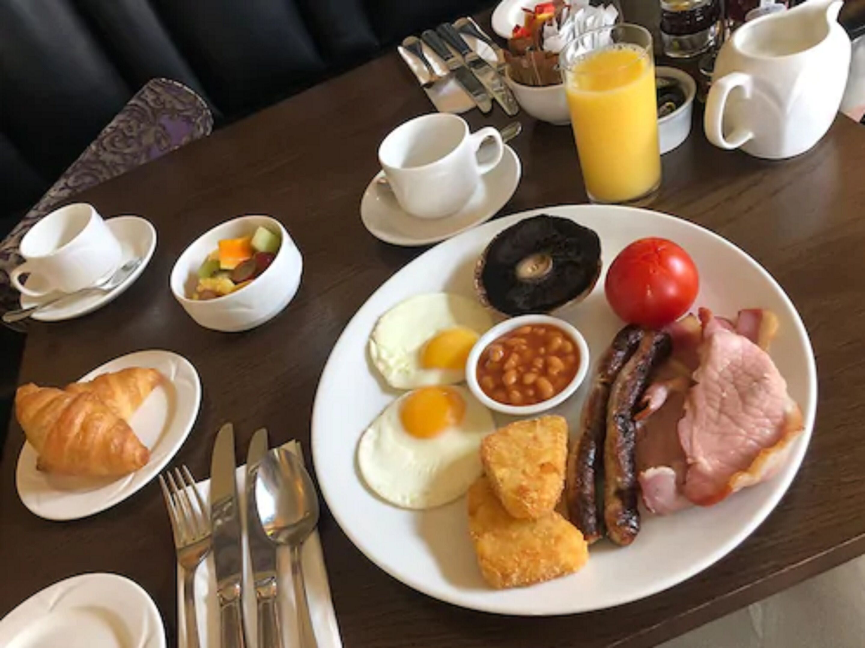 A breakfast table with plates of eggs, beans, sausages, ham, and a croissant. There are also cups, a jug of orange juice, and a bowl of fruit.