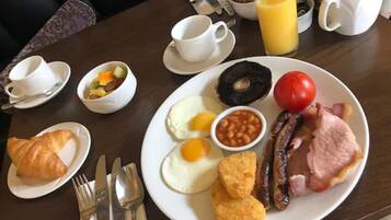 A breakfast table with plates of eggs, beans, sausages, ham, and a croissant. There are also cups, a jug of orange juice, and a bowl of fruit.
