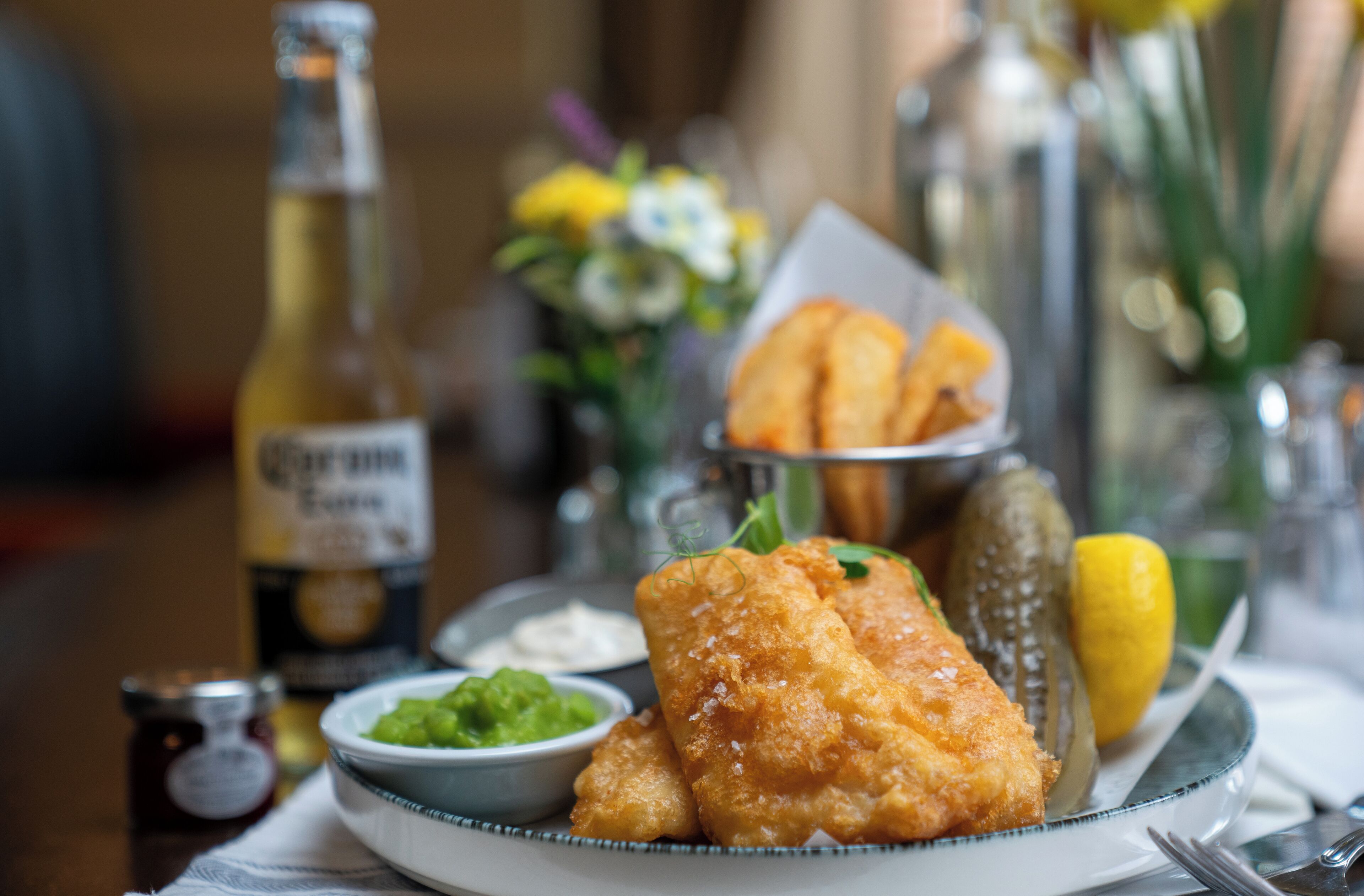 Un piatto di pesce fritto, limone e sottaceti su un tavolo con una bottiglia di birra e una piccola ciotola di salsa.