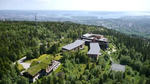 Complejo de edificios modernos rodeado de un bosque denso, con vistas a una ciudad distante y una masa de agua.