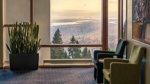 Habitación con un amplio ventanal que ofrece vistas a un paisaje urbano y montañas. Hay una planta en maceta y dos sillas.