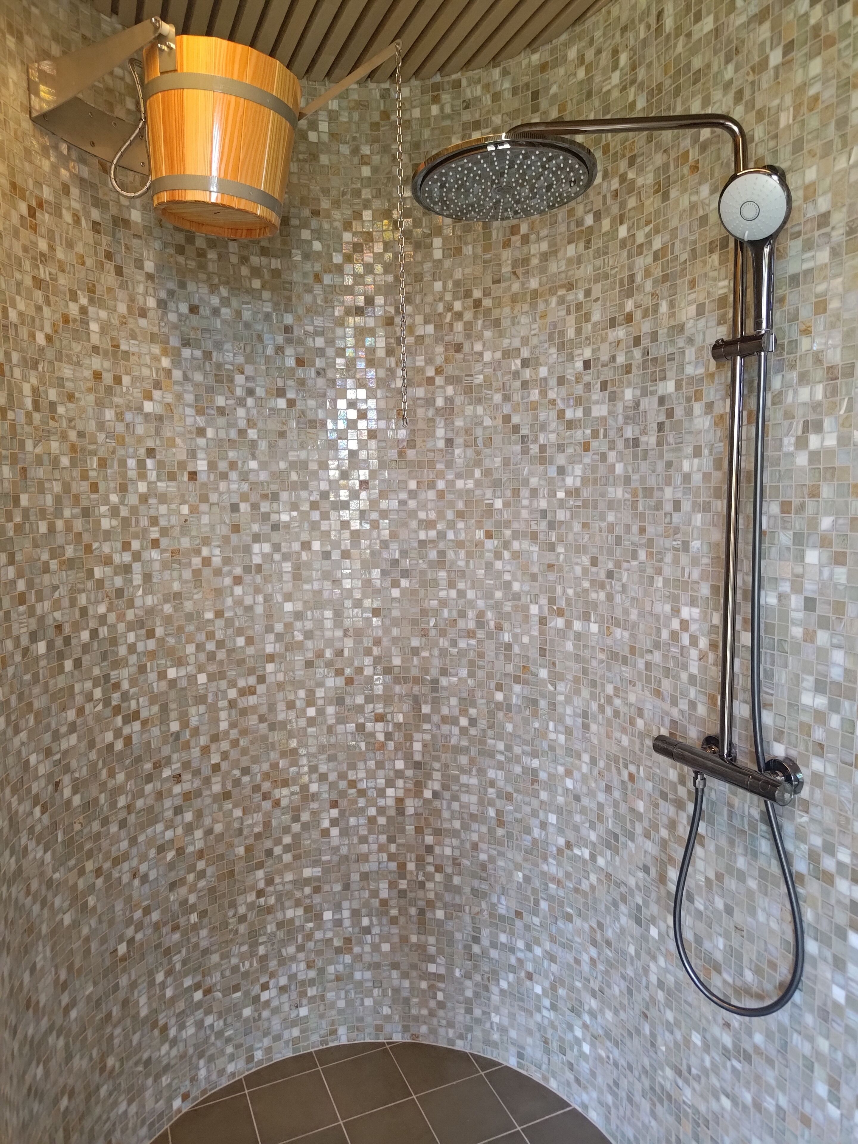 A shower with a mosaic tile wall, a wooden bucket, and a handheld showerhead.