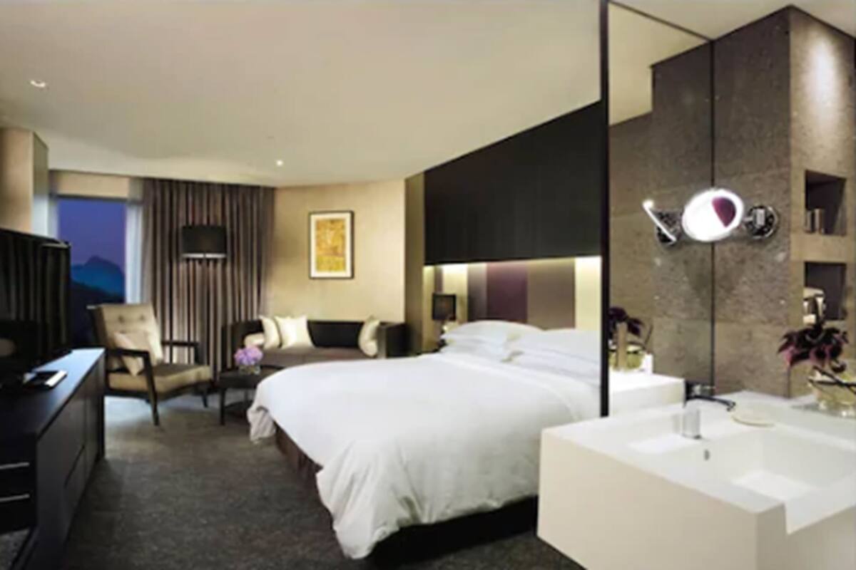A modern hotel room with a large bed, a desk, a chair, a large mirror, and a bathroom with a sink and mirror.