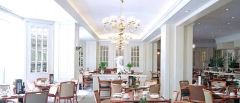 A spacious dining area with wooden tables and chairs, a central chandelier, and large windows allowing natural light.