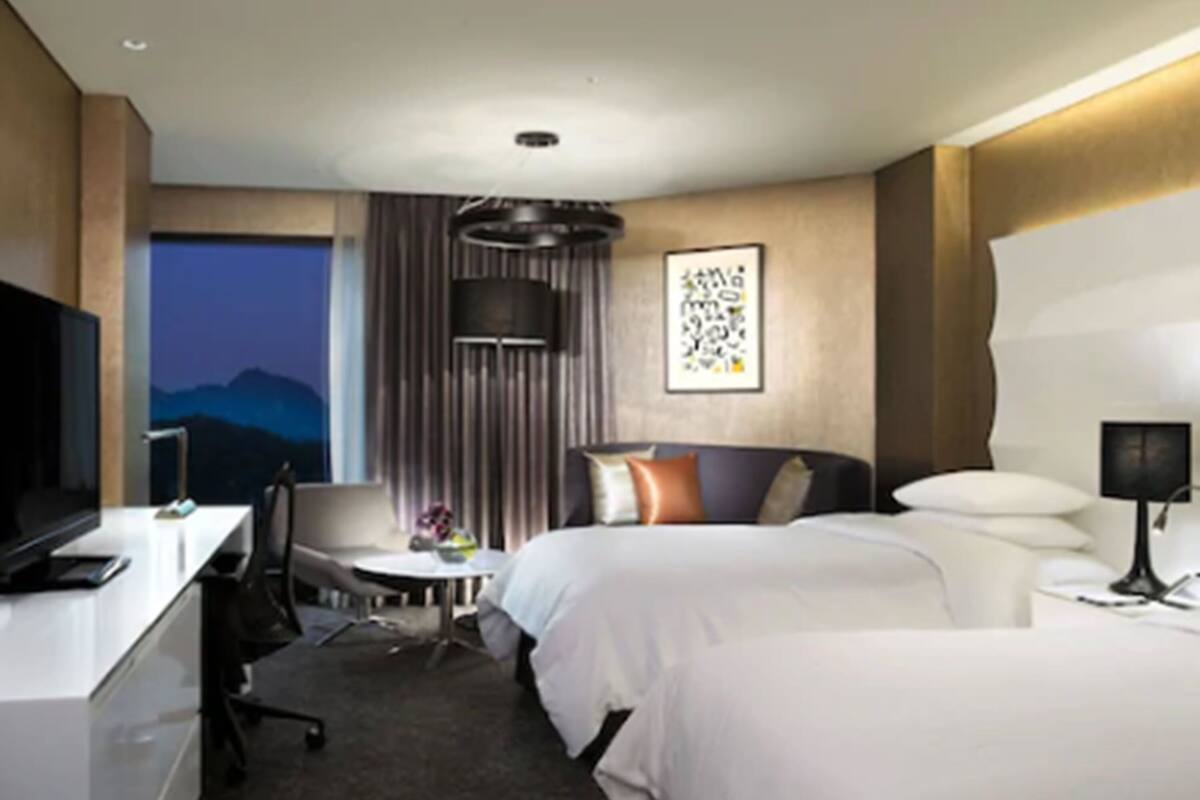 A hotel room with two beds, a desk, a chair, a TV, and a window with a view of mountains.