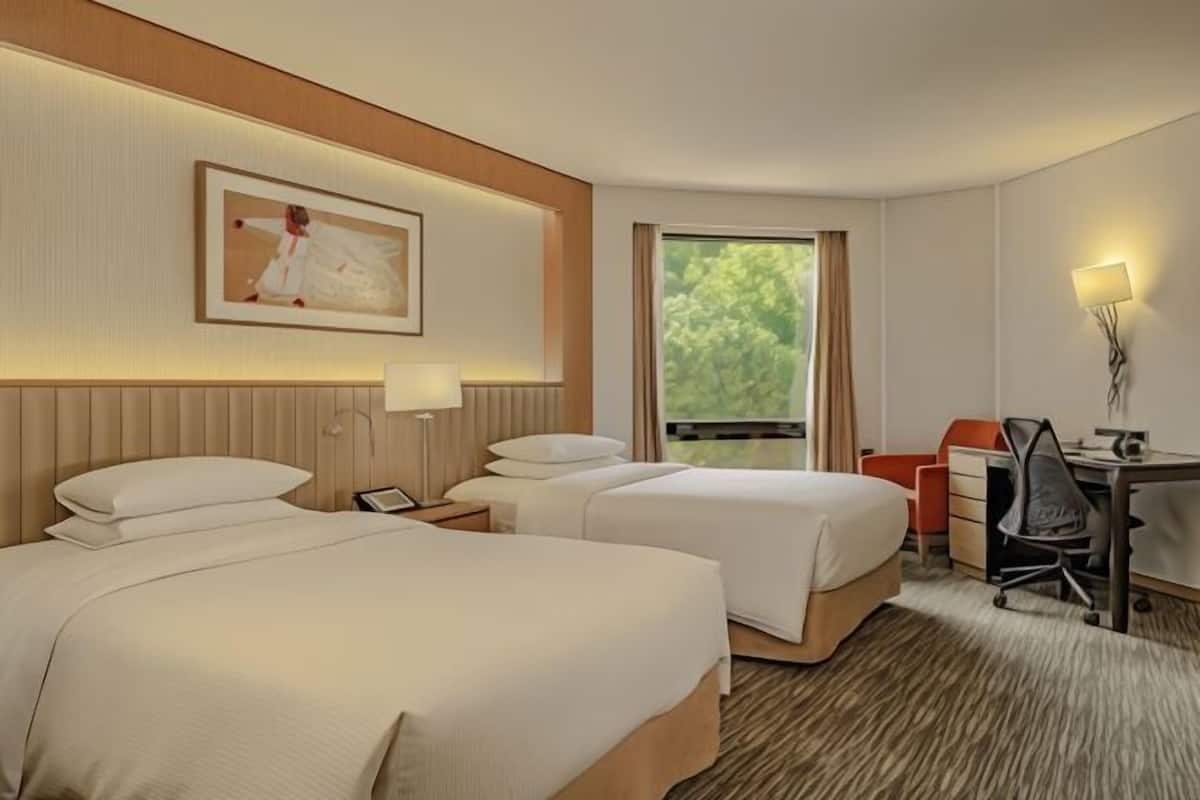 A hotel room with two beds, a desk, a chair, a lamp, and a window with a view of greenery.