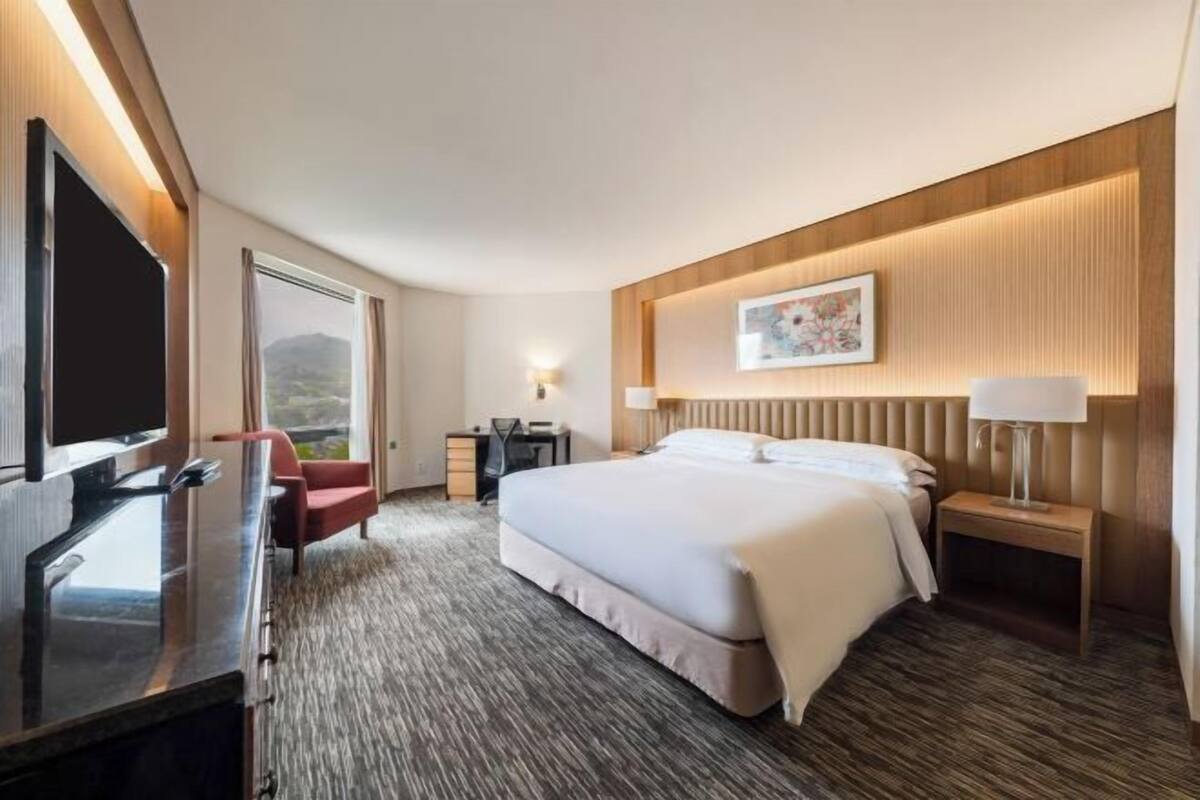 A hotel room with a bed, bedside tables, a desk, a TV, and a window with a view.