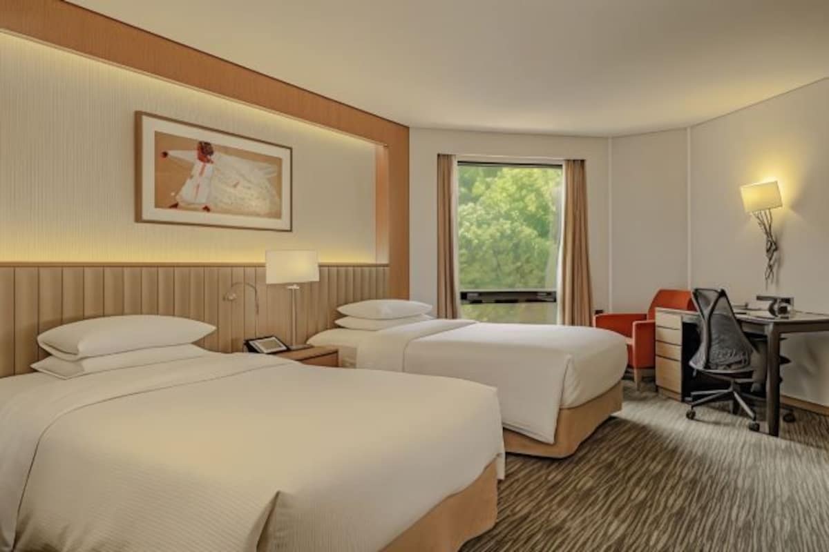 A hotel room with two beds, a desk, a chair, a lamp, and a window with a view of greenery.