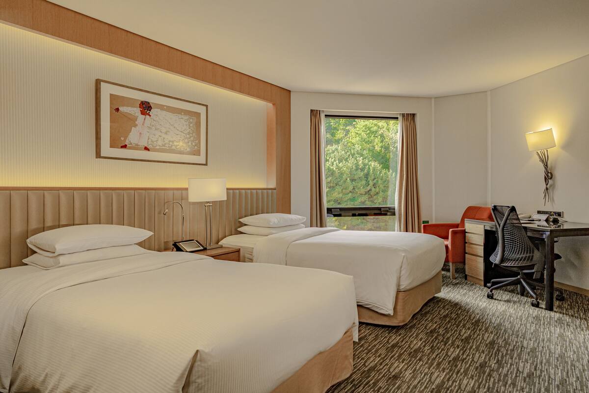 A hotel room with two beds, a desk, a chair, a lamp, and a window with a view of greenery.