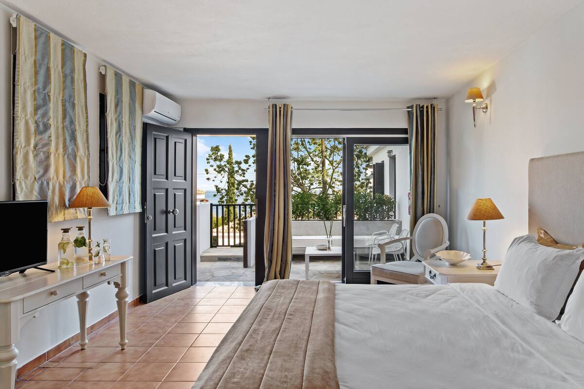 Habitación de hotel con cama, escritorio con televisor, silla, mesa con lámpara y balcón con vistas.