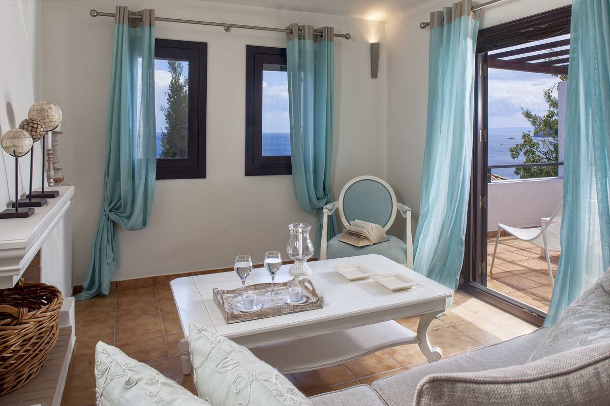 Una habitación con una mesa de comedor lista para dos personas, una silla, una ventana con cortinas y vista al mar.