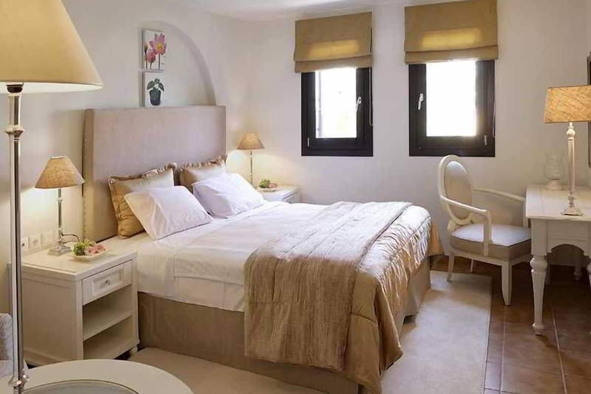 Un dormitorio con una cama, dos mesitas de noche con lámparas, un escritorio con silla, y dos ventanas con persianas.