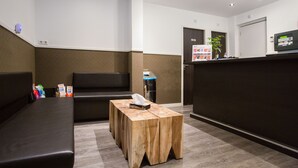 Zona de recepción con un banco de madera, un sofá negro, un cubo de basura y un mostrador de recepción con un monitor de computadora.