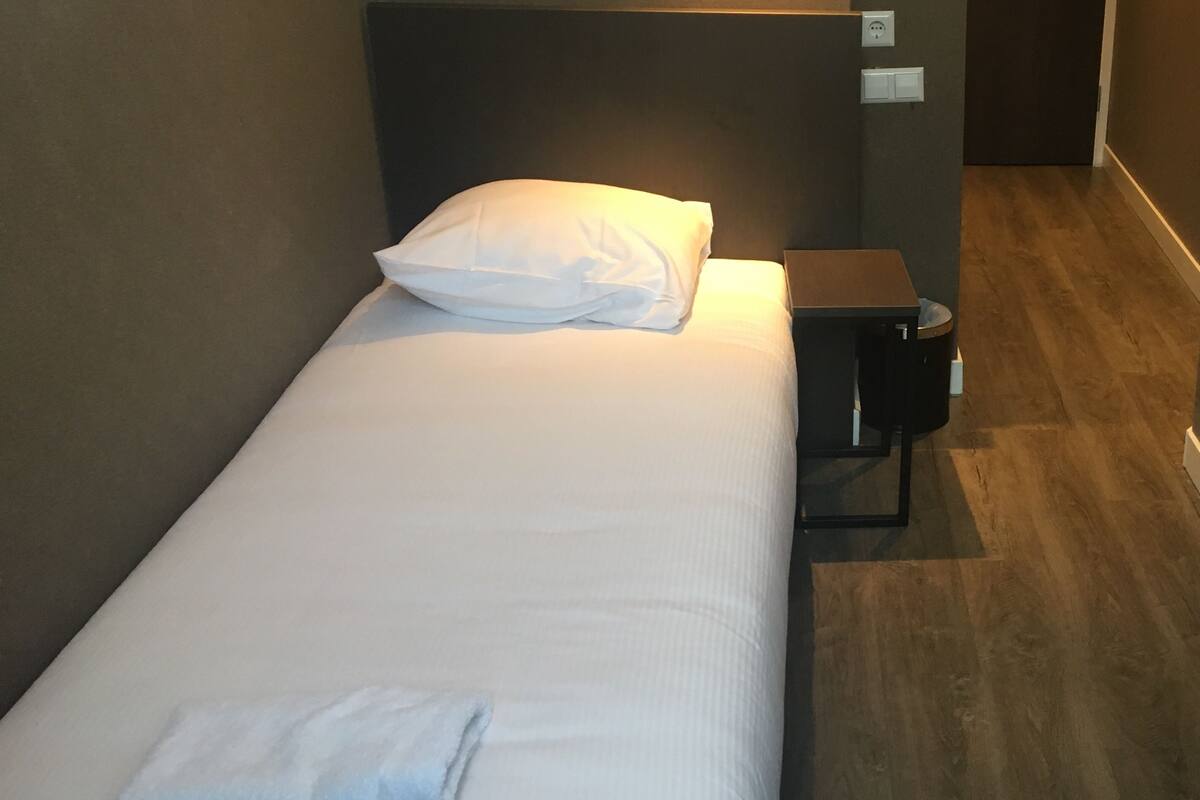 Una cama individual con ropa blanca, un almohadón y una toalla doblada. Hay una mesita de noche con una lámpara y un cubo de basura. El cuarto tiene piso de madera y un cabecero oscuro.