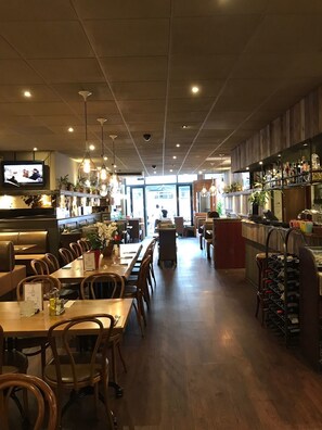 A restaurant with wooden tables and chairs, a bar area with bottles, and a television mounted on the wall. - Hotel Manofa (Amsterdam)
