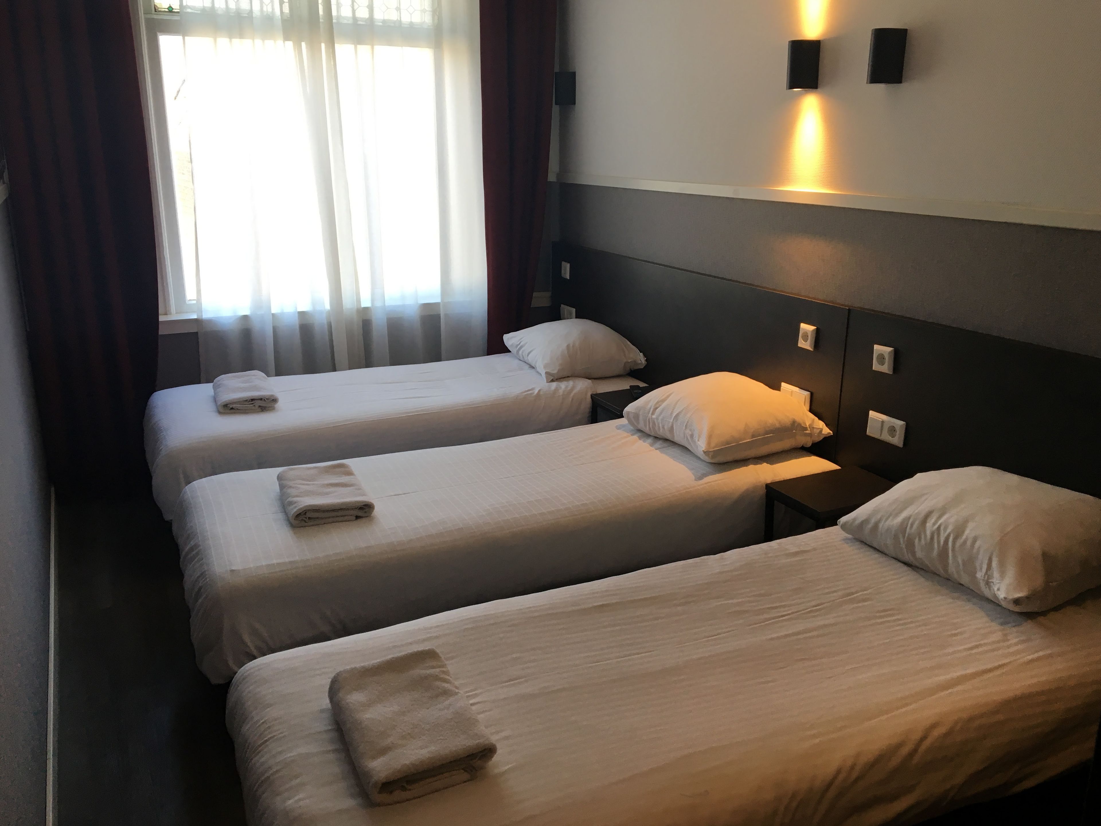 Une chambre d’hôtel avec trois lits simples, chacun recouvert d’une couette blanche et agrémenté d’une serviette pliée. Il y a des tables de chevet avec des lampes et des interrupteurs d’éclairage fixés au mur.