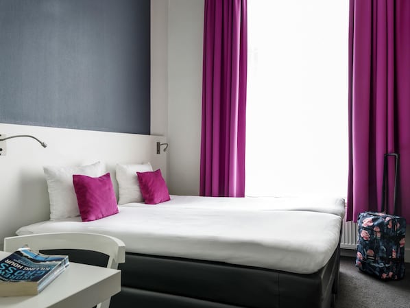 A hotel room with a bed, white and purple pillows, a white table with a book, a black chair, and a suitcase.