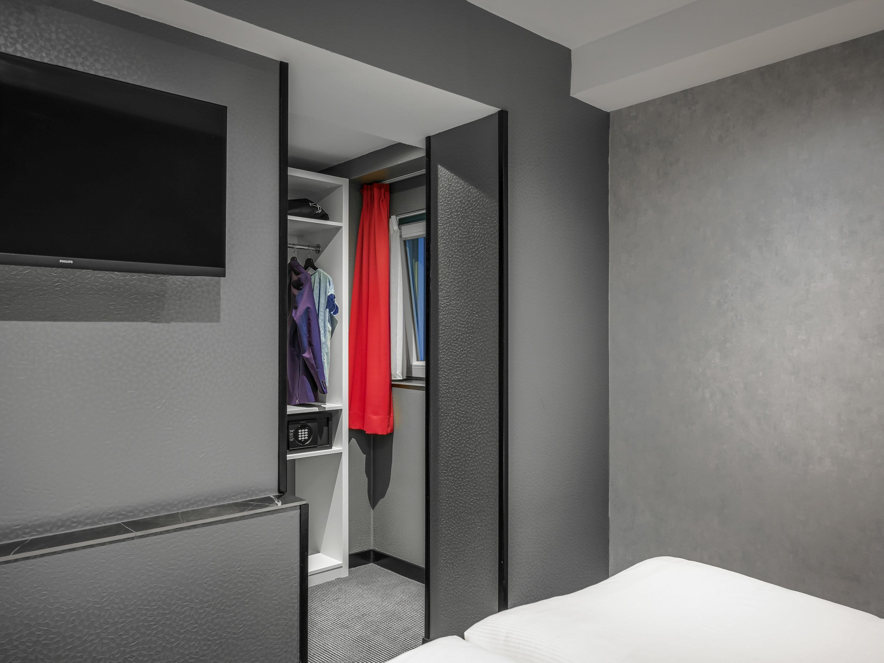 A hotel room with a bed, a television mounted on the wall, a closet with hanging clothes, and a red curtain.