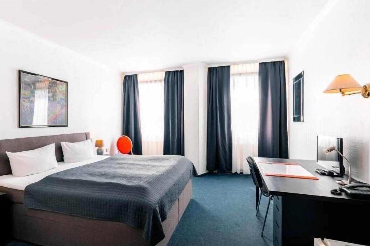 A hotel room with a bed, desk, chair, and a painting on the wall.
